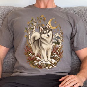 May include: A charcoal gray t-shirt featuring a graphic illustration of a husky standing on a snowy, rocky outcrop. The scene includes birch trees, pink heather flowers, a golden crescent moon, and distant mountains under a starry night sky.