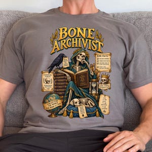 Könnte beinhalten: Ein graues T-Shirt mit einer Grafik eines Skeletts in einer blauen Robe, das ein Buch mit dem Titel All Things End Here liest. Das Design enthält einen Raben, einen Globus, ein Hundeskelett, eine Sanduhr und Schriftrollen mit Text wie Bone Archivist.