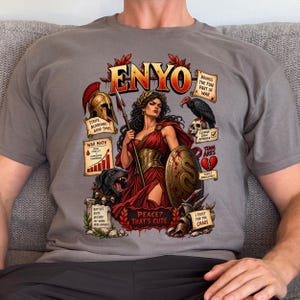 May include: A grey t-shirt featuring a graphic print of the goddess Enyo holding a spear and shield. The design includes a helmet, a black dog, a vulture, a skull, and text such as "ENYO," "WAR MATH," "PEACE? THAT'S CUTE," and "I EXIST FOR THE CHAOS."