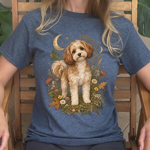 May include: A heather blue short-sleeve t-shirt featuring a central graphic of a fluffy tan and white dog standing amidst colorful wildflowers and autumn leaves, with a golden crescent moon and small stars positioned above the dog's head.