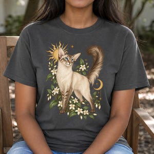 Peut inclure: Un t-shirt gris anthracite orné d'une illustration graphique représentant un chat crème aux yeux bleus, debout parmi des fleurs de jasmin blanches. Le motif comprend un soleil doré, un croissant de lune et de petites étoiles.