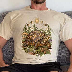 Könnte beinhalten: Ein beigefarbenes T-Shirt mit einer zentralen Grafik einer Dosenschildkröte auf einem moosigen Stein, umgeben von Farnen, Pilzen, Eicheln und weißen Blüten. Über der Schildkröte sind eine Sonne und Mondphasen kreisförmig dargestellt.