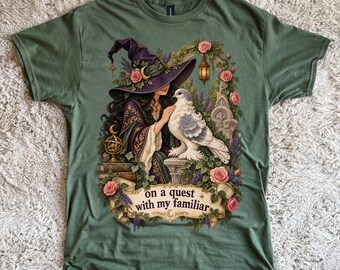 May include: An olive green t-shirt featuring a graphic print of a person in a purple witch hat and robe, touching a white dove. The design includes pink roses, a lantern, books, and a banner with the text, on a quest with my familiar.