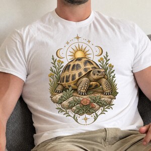 Pode incluir: Uma t-shirt branca com um gráfico central de uma tartaruga castanha sobre rochas, rodeada por folhagem verde e pequenas flores vermelhas. Acima da tartaruga está um sol dourado ladeado por duas luas crescentes e estrelas.