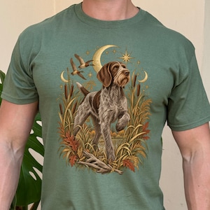 May include: A sage green short-sleeved t-shirt featuring a graphic illustration of a brown and white wirehaired pointing dog standing in tall grass and cattails. The scene includes flying ducks, a crescent moon, and stars in a golden hue.