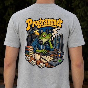 May include: A heather gray t-shirt featuring a back graphic of a green frog wearing a blue hoodie and headphones, sitting at a desk with a computer, books, a coffee mug, and a drink can. The text "Programmer" is printed in yellow above the frog.
