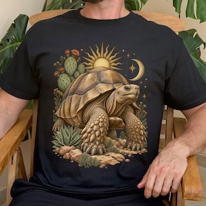 Op de afbeelding: Een wit t-shirt met een gedetailleerde afbeelding van een bruine schildpad op een rotsachtige ondergrond. Het ontwerp bevat een groene cactus, een gele zon, een maansikkel en kleine sterren, wat een woestijnachtig tafereel vormt.