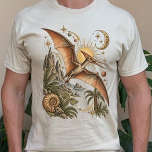 May include: A cream-coloured t-shirt featuring a detailed graphic of a flying pterodactyl soaring above a prehistoric landscape with palm trees, rocky cliffs, a large ammonite fossil, a golden sun, a crescent moon, and various celestial stars.