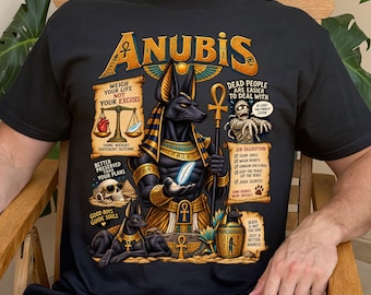 May include: A black t-shirt featuring a graphic of the Egyptian deity Anubis holding an ankh staff and a feather. The design includes text such as 'ANUBIS', 'WEIGH YOUR LIFE NOT YOUR EXCUSES', 'DEAD PEOPLE ARE EASIER TO DEAL WITH', and a job description list.