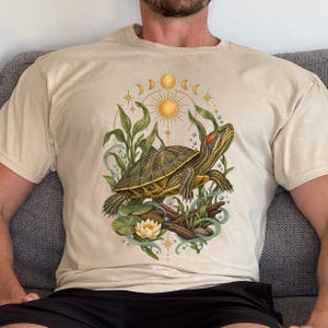 Op de afbeelding: Een beige t-shirt met een grafische illustratie van een roodwangschildpad die zwemt tussen groene waterplanten, leliebladeren en een witte waterlelie, omlijst door een gouden zon, maanfasen en kleine sterren in een cirkelvormig ontwerp.