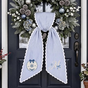 Può includere: Un appendiporta in tessuto a righe blu e bianche con bordi smerlati, caratterizzato da una ghirlanda di fiocchi di neve ricamata su un lato e una lettera M monogrammata con un fiocco blu sull'altro, esposto su una ghirlanda di pino.