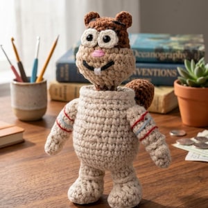 Puede incluir: Una figura de ardilla tejida a ganchillo que lleva un traje espacial beige con rayas rojas y azules en los brazos. La figura tiene una cabeza marrón, ojos grandes blancos con pupilas negras, una nariz rosa y dos dientes blancos.