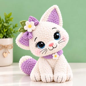 May include: A white crocheted cat plush toy with large blue eyes, a pink nose, and long eyelashes. It features a purple bow with a white flower and green leaves on its head, a purple collar with a small white flower, and a purple-tipped tail.