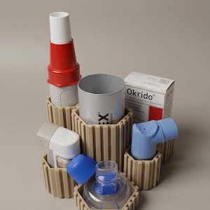 Może przedstawiać: Beżowy organizer z wieloma przegródkami mieści różne inhalatory medyczne oraz pudełko leku Okrido. Organizer ma żebrowaną, cylindryczną konstrukcję z kilkoma oddzielnymi otworami, w których znajdują się czerwono-biały inhalator, niebieski inhalator oraz przezroczysta maska.