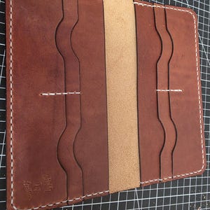 May include: A brown leather bifold wallet featuring white contrast stitching, multiple card slots with wavy edges, and a central cash compartment. The leather is embossed with the text Muddy Dog Leather in the bottom left corner of the wallet.