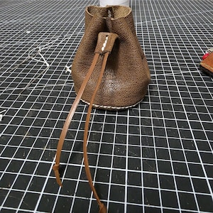 May include: A brown textured leather drawstring pouch featuring a circular base with visible white stitching along the bottom edge. The pouch has a leather cord closure that extends downward, and the top opening is secured with a leather loop.