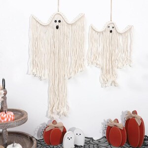 May include: Two off-white macrame ghost wall hangings with black button eyes, suspended by thin cords. Below them are several small wooden pumpkins in shades of red and orange, along with two small white ghost figures on a black lace surface.