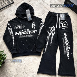 May include: A black hooded sweatshirt and matching flared sweatpants set featuring white graphic prints. The hoodie displays the text Hellstar and Faith to Fairydise with flame motifs. The pants include white drawstrings and matching branding.