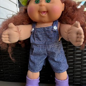 May include: A doll with curly reddish-brown hair, green eyes, and rosy cheeks, wearing a lime green and lavender beret, blue denim overalls with white buttons, and lavender rubber boots. The doll has a smiling expression with one visible tooth.