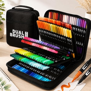 Könnte beinhalten: Eine schwarze Transporttasche mit einem großen Set an Dual-Brush-Stiften in einer breiten Farbpalette. Die Tasche trägt den weißen Schriftzug DUAL BRUSH PEN. Die Stifte sind in gestuften Fächern angeordnet und zeigen verschiedene Rot-, Orange-, Gelb-, Grün-, Blau- und Violetttöne. Zwei Stifte liegen außerhalb der Tasche, einer mit orangefarbener und einer mit schwarzer Spitze, auf weißem Papier mit orangefarbenen Tintenstrichen.