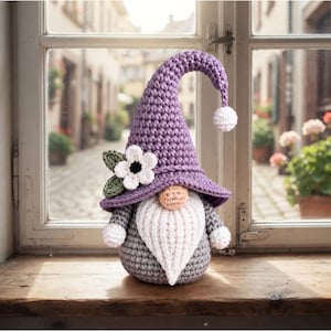 May include: A crochet gnome figurine featuring a tall, pointed purple hat with a white pom-pom at the tip, adorned with a white flower and green leaves. The gnome has a round peach-colored nose, a long white beard, and a gray rounded body.