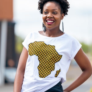 May include: A woman wearing a white t-shirt with a yellow and brown graphic of the continent of Africa. She is smiling and looking to the right. She is wearing black jeans and has short, curly black hair.