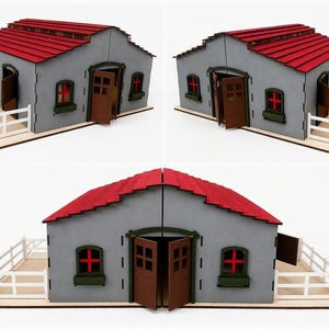 May include: A model horse stable featuring a grey structure with a red roof, brown doors, and green window frames. The building includes white fencing on both sides, with open doors and windows showing red cross details on the interior panels.