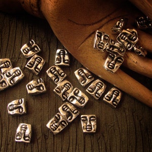 May include: A collection of silver-toned metal beads with a face design. The beads are scattered on a brown fabric surface.