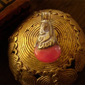 May include: A silver pendant with a pink stone set in a decorative silver frame. The pendant is resting on a gold-colored surface with intricate designs.