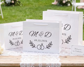 Op de afbeelding: Drie witte papieren cadeauzakjes met witte koordhengsels, elk voorzien van zwarte opdruk M & D, de datum 09.28.2026 en een afbeelding van twee in elkaar grijpende ringen met een bladmotief. Een klein wit kaartje bevat de tekst Welcome to our Celebration.