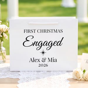May include: A white rectangular gift bag with white cord handles. The front features black text reading FIRST CHRISTMAS Engaged, a small star icon, the names Alex & Mia, and the year 2026 printed in a combination of serif and script fonts.