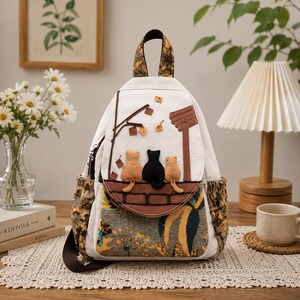 Pode incluir: Uma mochila de lona branca com uma aba apresentando uma aplicação de três gatos sentados numa cerca castanha debaixo de uma árvore. A mala tem uma base e bolsos laterais de tecido com um padrão floral amarelo, castanho e azul.