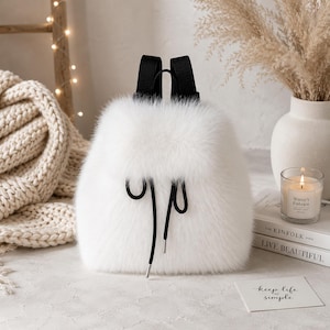 Puede incluir: Una mochila de piel sintética blanca con una textura suave y esponjosa, correas negras ajustables para los hombros y un cierre de cordón negro con puntas de metal plateado. El bolso tiene una forma redondeada y un exterior denso y blanco.