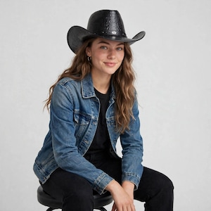 May include: A black cowboy hat with a textured crocodile skin pattern and a thin band featuring a small silver-toned metal accent. The hat has a wide, curved brim and a high crown, constructed from a dark material with a glossy finish.