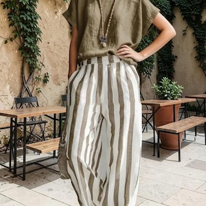 Puede incluir: Una blusa de manga corta color verde oliva combinada con pantalones de tiro alto y pernera ancha con rayas verticales en verde oliva y blanco. El conjunto se complementa con un largo collar de cuentas con un colgante central, creando un estilo coordinado.