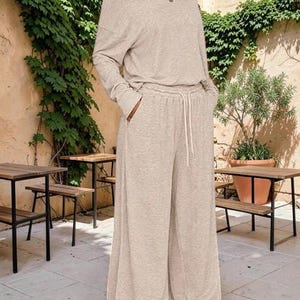 May include: A beige two-piece lounge set consisting of a long-sleeved top with a wide neckline and matching wide-leg trousers. The trousers feature an elasticated waistband with a drawstring and side pockets, crafted from a soft, heathered fabric.