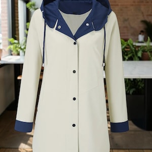May include: A long-sleeved raincoat in a light cream color featuring a contrasting dark navy blue hood and matching cuffs. The coat has a front button closure with dark navy buttons, a drawstring hood, and two side pockets on the front panel.