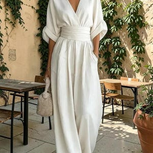 Könnte beinhalten: Ein einfarbiger weißer Jumpsuit mit tiefem V-Ausschnitt, ellbogenlangen Ärmeln mit umgeschlagenen Bündchen und einem breiten, gerafften Taillenbund. Das Kleidungsstück hat eine weite Hose mit Seitentaschen und eine glatte, matte Stoffoberfläche.