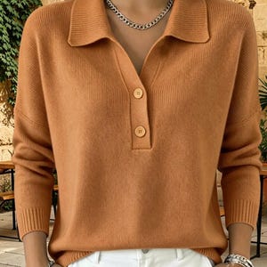 Può includere: Un maglione in maglia color marrone chiaro a maniche lunghe con colletto e un'abbottonatura parziale con due bottoni visibili. Il capo presenta polsini e orlo a coste, mettendo in risalto un tessuto in maglia morbido e testurizzato.