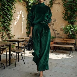 May include: A dark emerald green velvet two-piece set consisting of a long-sleeved mock neck top and wide-leg trousers. The fabric has a soft, reflective sheen. A black handbag is held at the side, and light-coloured pointed-toe shoes are visible.