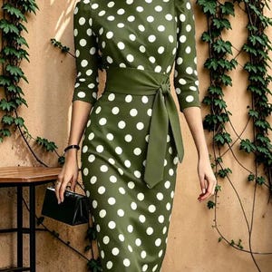 May include: A midi-length olive green dress featuring a white polka dot pattern, three-quarter length sleeves, a round neckline, and a matching fabric tie-waist belt. The garment has a fitted silhouette and is paired with a small black handbag.