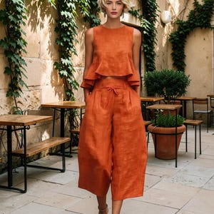 Op de afbeelding: Een tweedelige set bestaande uit een mouwloze, cropped peplumtop en een wijde culottebroek in een effen roestoranje kleur. De broek heeft een hoge taille met een trekkoord in het midden en twee zijzakken.