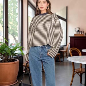 Puede incluir: Una camiseta de manga larga con rayas horizontales marrones y blancas y un cuello oscuro sólido, combinada con pantalones vaqueros azules. La camiseta tiene un ajuste relajado con hombros caídos, mientras que los vaqueros tienen un corte recto clásico.