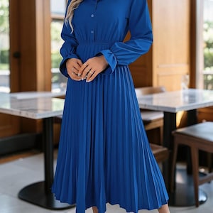 Könnte beinhalten: Ein königsblaues Midikleid mit Hemdkragen, durchgehender Knopfleiste am Oberteil und langen Ärmeln mit gerüschten Manschetten. Das Kleid hat eine betonte Taille und einen weit ausgestellten, plissierten Rock, der unterhalb der Knie endet.
