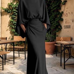 May include: A floor-length black dress featuring a boat neckline, wide cape-style sleeves, and a gathered waistline. The garment has a smooth, flowing silhouette that extends to the floor, creating a clean and structured appearance throughout.