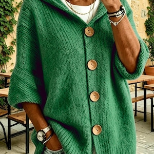 May include: A green hooded cardigan featuring a textured knit pattern and four large, round, light brown wooden buttons down the front. The garment has three-quarter length sleeves and is worn over a plain white shirt with light blue denim jeans.