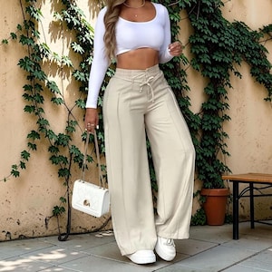 May include: A long-sleeve white cropped top with a scoop neckline paired with high-waisted beige wide-leg trousers featuring a front drawstring tie and vertical seam detailing, accompanied by a white quilted shoulder bag with a gold chain strap.