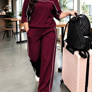 May include: A burgundy two-piece lounge set consisting of a short-sleeved top and wide-leg trousers, both featuring thin white piping accents. The outfit is paired with white trainers, a black quilted backpack, and a light pink rolling suitcase.