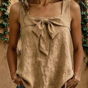 May include: A sleeveless tan tank top featuring a square neckline and a prominent fabric bow detail centered at the chest. The garment is crafted from a textured material with a subtle crinkled finish, providing a structured yet relaxed fit.