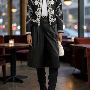 Könnte beinhalten: Eine schwarze Jacke mit weißer Blumenstickerei und Rundhalsausschnitt, getragen über einem weißen Oberteil. Das Outfit umfasst einen schwarzen Hosenrock in Lederoptik mit goldfarbener Gürtelschnalle sowie schwarze kniehohe Wildlederstiefel mit spitzer Kappe und Stilettoabsatz.